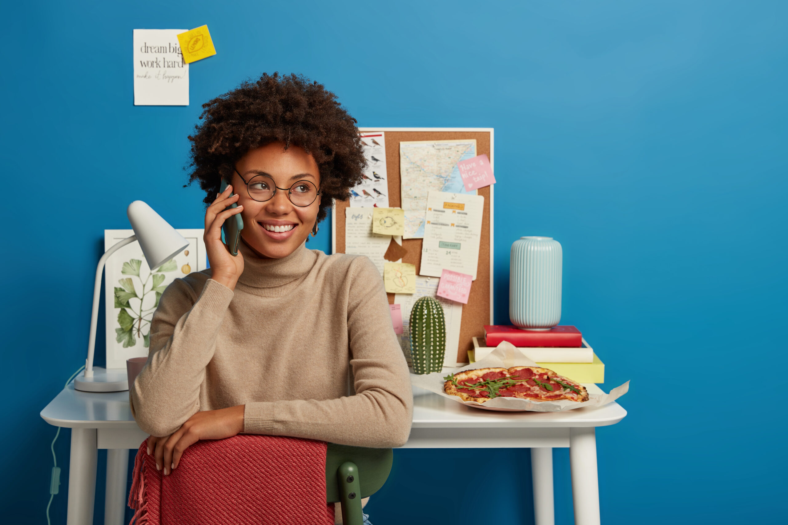 Cheerful curly woman has corporate communication via cellphone, looks aside with smile, poses in coworking space, eats pizza during break. Happy student talks with groupmate, discuss exam preparation